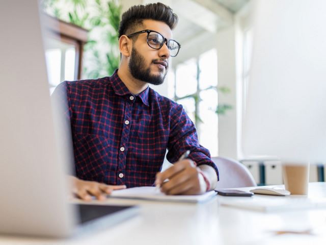 Man writing in notebook and looking at computer monitor