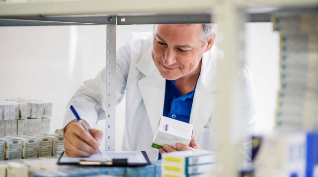 ein Apotheker schreibt Notizen auf einen Block, während er eine Bestandsaufnahme in einem Regal mit verschiedenen Medikamenten durchführt