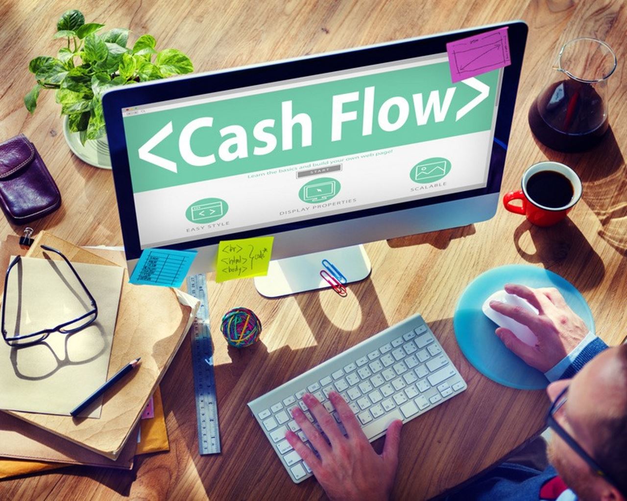 man working on desktop computer with Cash Flow wallpaper surrounded by office supplies and coffee