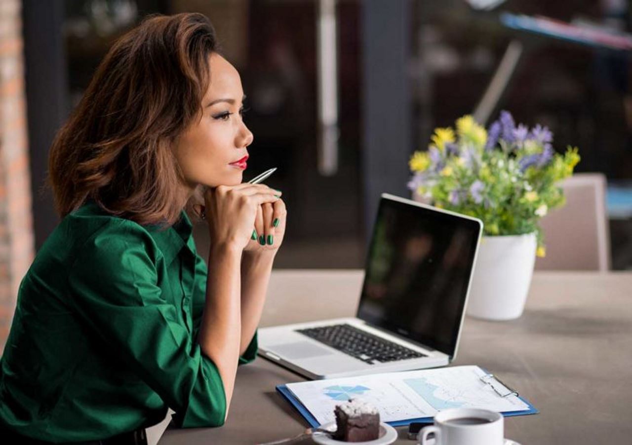 woman sitting at table with laptop and clipboard, holding pen and thinking